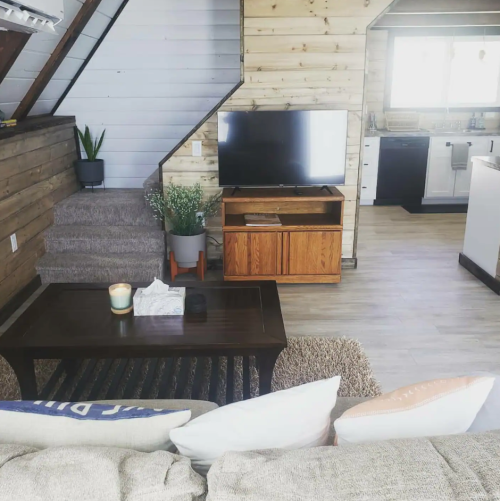 Cozy living room with a sofa, coffee table, TV, and open kitchen, featuring wooden walls and a staircase.